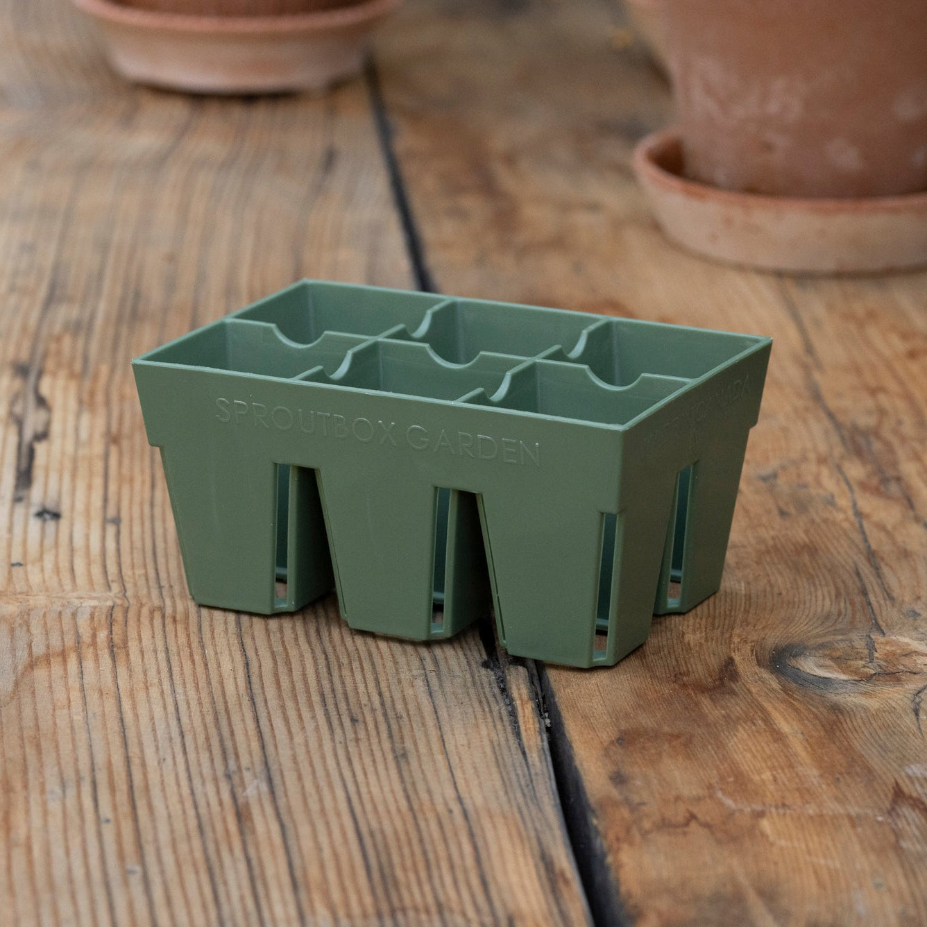 Green plant potting tray on a wooden surface with terracotta pots in the background